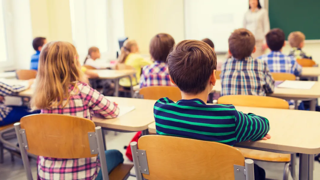 Image of the back of a classroom with several children sitting at their desks facing their teacher and a blackboard.