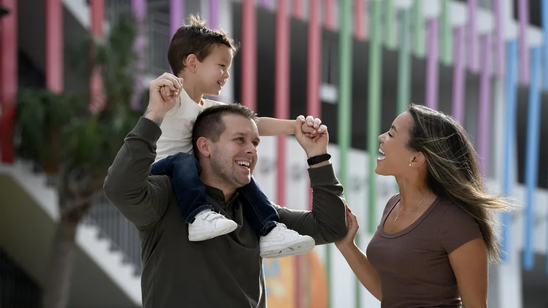 A child riding on his fathers shoulders while his smiling mother walks along side.