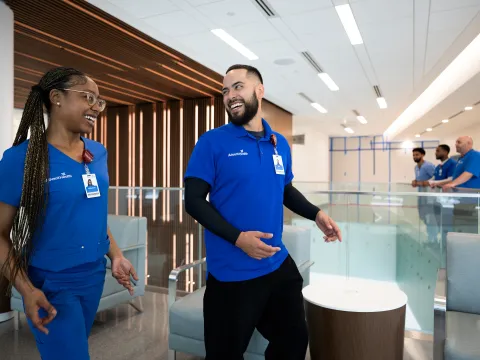 Two AdventHealth team members, a woman and a man, smile and walk together through a bright, modern healthcare facility with contemporary lighting and furniture. Both wear blue uniforms with visible ID badges. In the background, three other colleagues in similar attire are conversing near a glass railing on the upper level.
