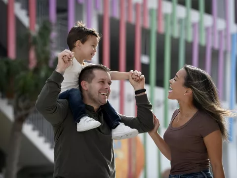 A child riding on his fathers shoulders while his smiling mother walks along side.