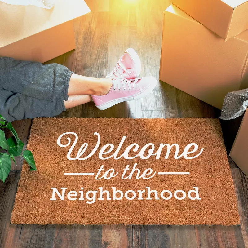 Person unpacking and a door mat with "Welcome to the Neighborhood" printed on it sits on the floor.