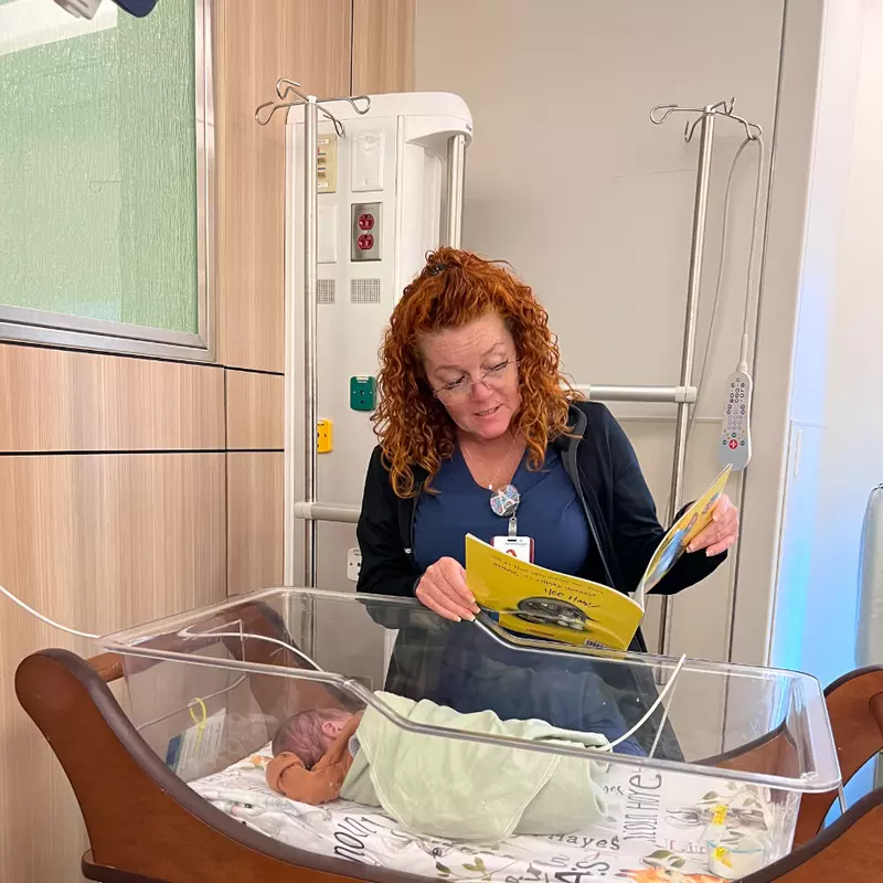 April Nelson, a nurse at AdventHealth for Children, stands over a crib, reading to a swaddled baby in the NICU.