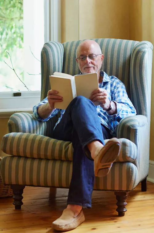 An older man reading a book while seating on a chair.